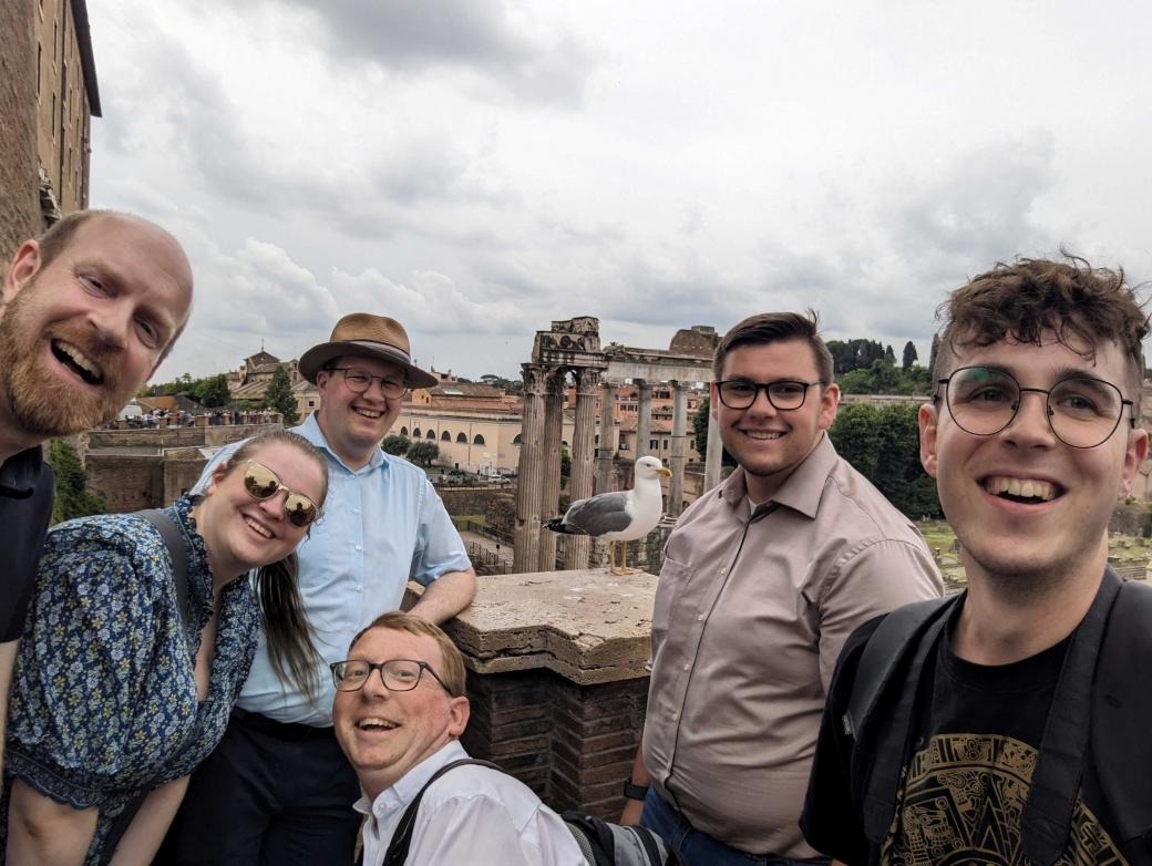 sechs Personen machen ein Selfie. Im Hintergrund sieht man Gebäude aus dem alten Rom