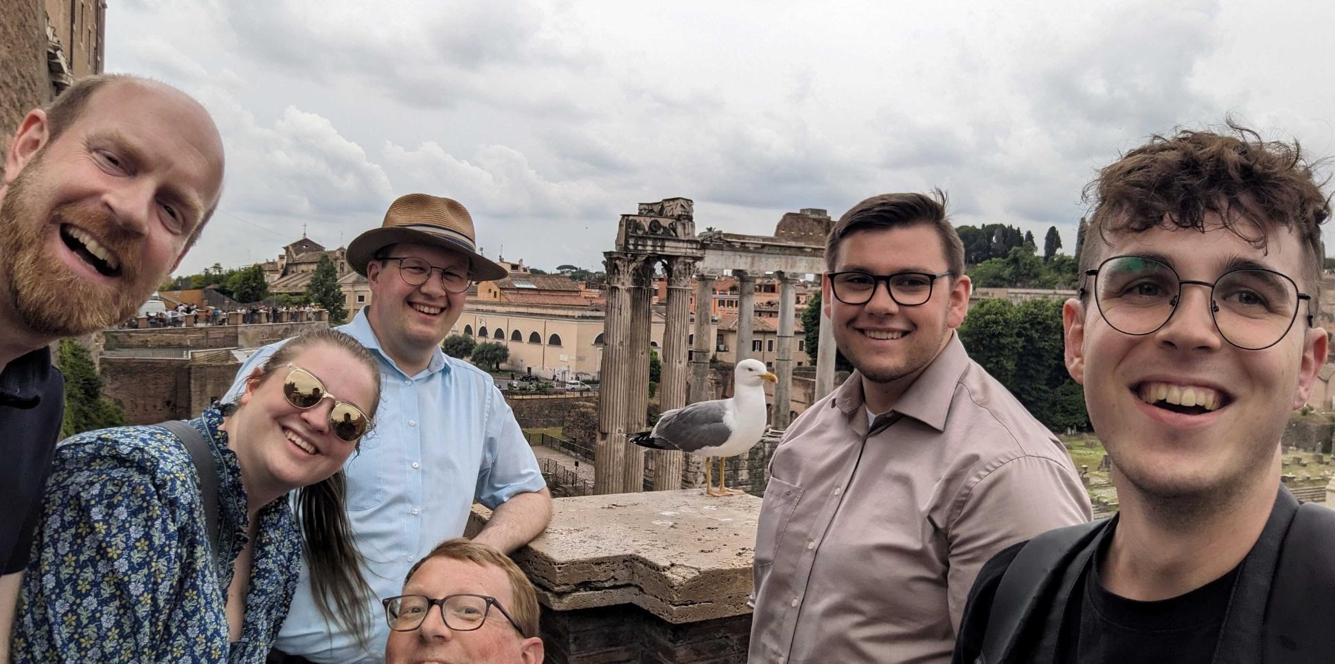 sechs Personen machen ein Selfie. Im Hintergrund sieht man Gebäude aus dem alten Rom