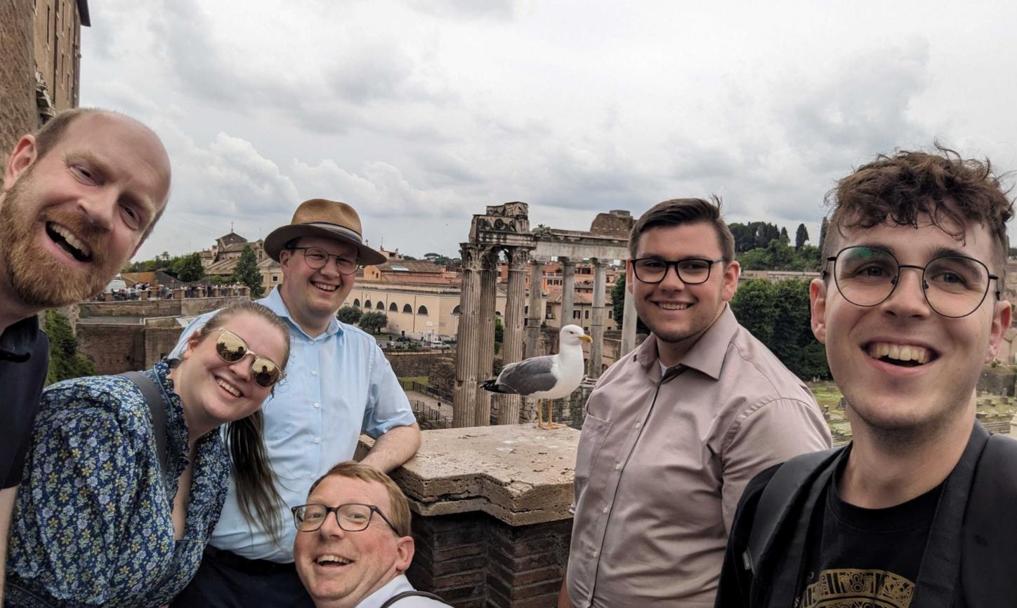 sechs Personen machen ein Selfie. Im Hintergrund sieht man Gebäude aus dem alten Rom