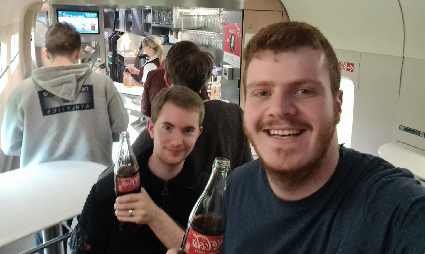 Zwei Teilnehmende im Boardbistro der Bahn mit einem Getränk in der Hand