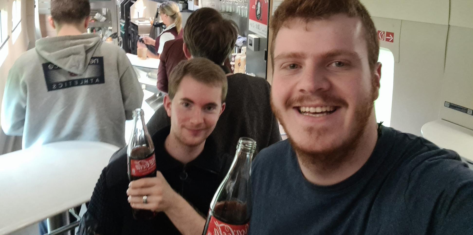 Zwei Teilnehmende im Boardbistro der Bahn mit einem Getränk in der Hand