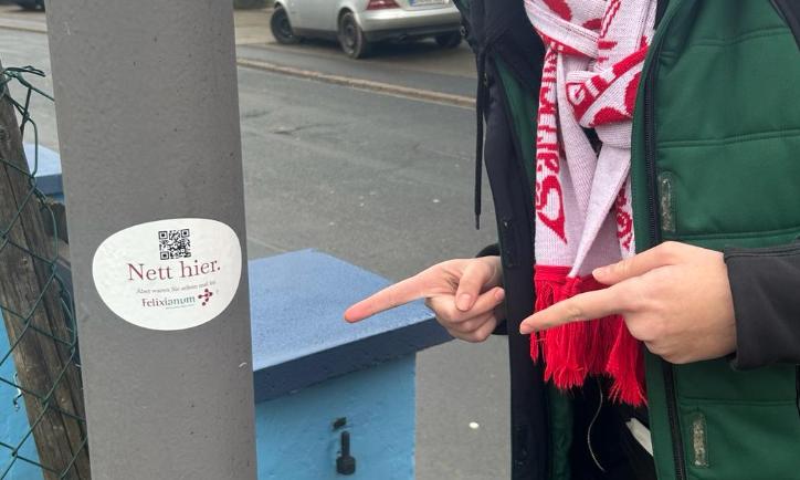 Mensch zeigt mit zwei Fingern auf einem Laternenpfahl. Dort klebt ein Aufkleber mit der Aufschrift Nett hier, aber waren sie schon im Felixianum?