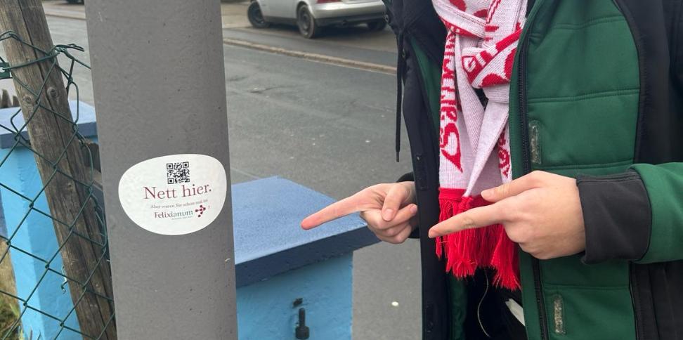 Mensch zeigt mit zwei Fingern auf einem Laternenpfahl. Dort klebt ein Aufkleber mit der Aufschrift Nett hier, aber waren sie schon im Felixianum?