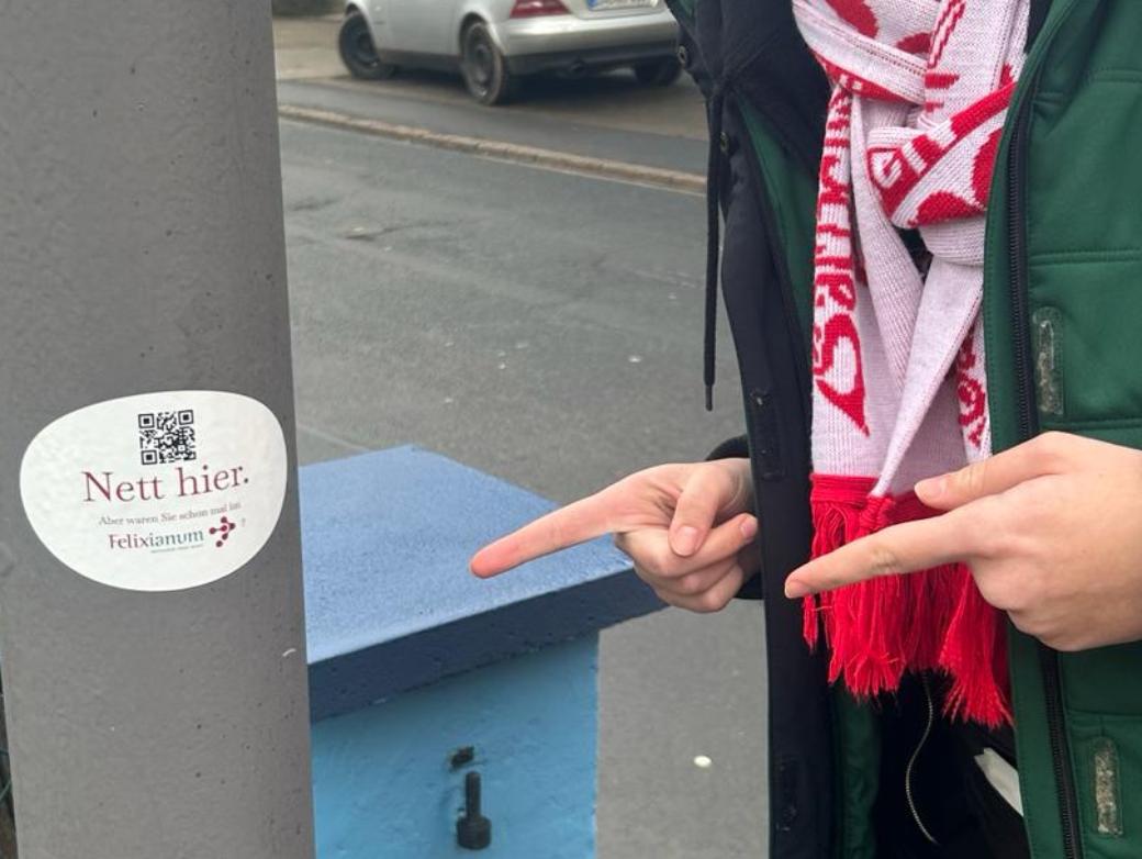 Mensch zeigt mit zwei Fingern auf einem Laternenpfahl. Dort klebt ein Aufkleber mit der Aufschrift Nett hier, aber waren sie schon im Felixianum?